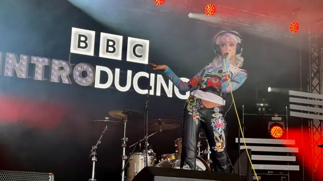A woman stood on stage with headphones on singing into a mic with a yellow cord attached to it. Behind her are the words BBC Introducing on a black background
