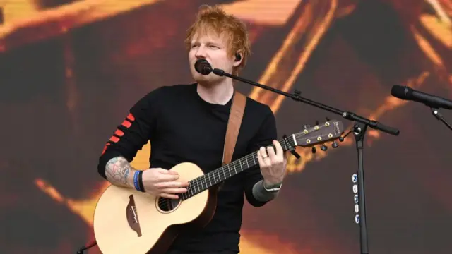Ed Sheeran sings into a microphone while playing a guitar at Big Weekend in 2022. He is wearing a black, long-sleeved t-shirt and he has blue eyes and ginger hair.