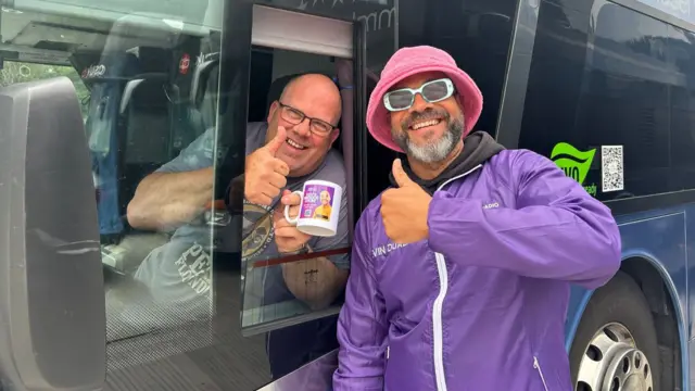 Kevin Duala and Rob give a thumbs up to the camera. Rob holds a BBC Radio Merseyside mug. Kevin is wearing a purple BBC Local Radio jacket, a pink bucket hat, and green sunglasses. Rob is wearing a grey graphic t-shirt.