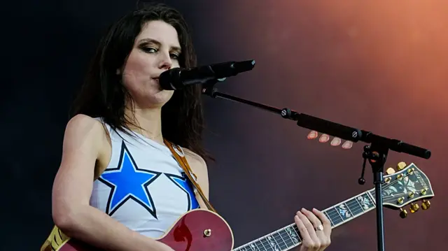 Ellie Rowsell sings into a microphone while playing a red guitar. She has long brown hair and is wearing a sleeveless white top with two blue stars on it.