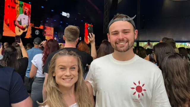 Blonde woman and man stand side by side as ed sheeran performs behind them, a crowd observing