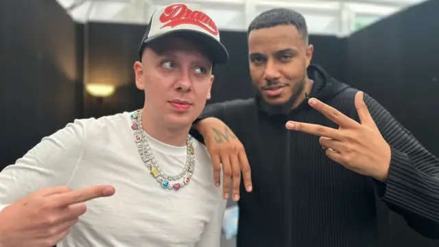 Aitch and AJ Tracey pose for the camera. Aitch is pointing at AJ Tracey while AJ Tracey holds up three fingers and leans on Aitch's shoulder. Aitch is wearing a white t-shirt, a silver necklace with colourful stones in it, and a baseball cap. He has blue eyes. AJ Trace is wearing a black jumper and has brown hair and brown eyes.