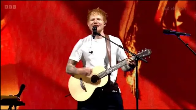 Ed Sheeran, wearing a white T-shirt, sings on stage and holds a guitar
