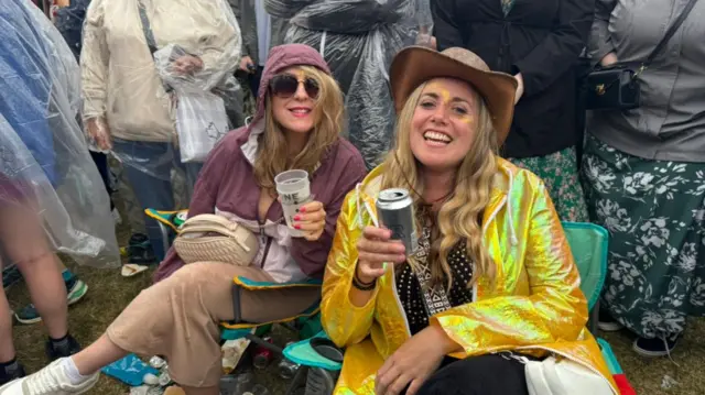 Rebecca and Becky smile for the camera while sat on green camping chairs and holding a drink each. Rebecca is wearing a maroon coat, beige trousers, white trainers, and sunglasses. Becky is wearing a shiny yellow jacket, a black and white studded top, black trousers, a brown cowboy hat, and yellow glitter on her face. Both women have blonde hair.