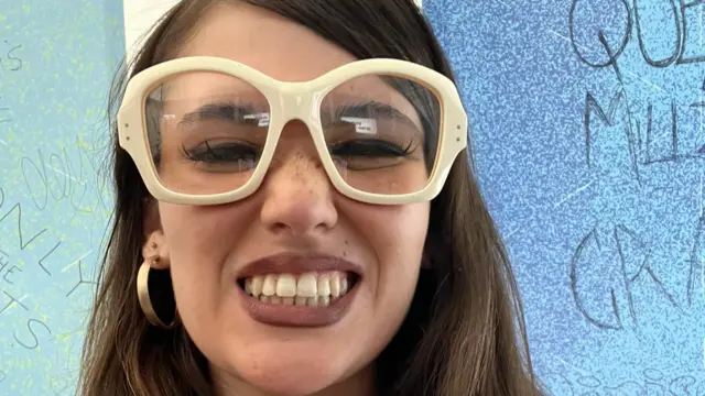 Sienna Spiro smiles for the camera. She is wearing white thick-framed glasses. She has long brown hair.
