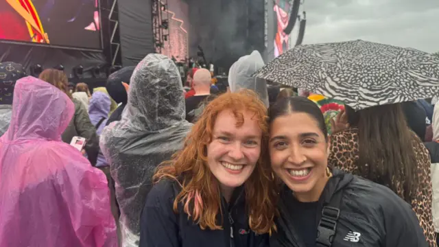 Hope and Mimi smile for the camera while stood in a crowd in front of a stage at Big Weekend. Hope has ginger hair and blue eyes and Mimi of them has brown eyes and brown hair. They are both wearing black coats while people around the are wearing poncho raincoats and one person is holding an umbrella.