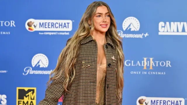 Charlotte Plank wears a black and brown checked coat at the MTV EMAs in 2024. She has blue eyes and long brown hair with blonde highlights in it.