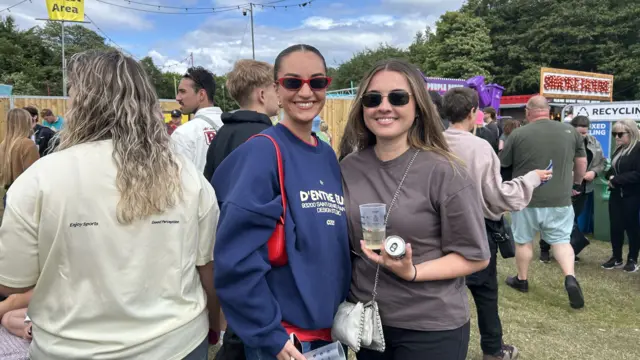 Best friends Anna Griffith and Holly Quinn smile at the camera. Anna, on the left, is wearing sunglasses and carrying a red purse. Holly is also wearing sunglasses and carrying a white bag and holding a drink.