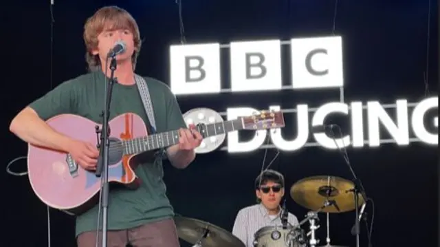 A man playing a guitar singing into a mic stand as a guy drums behind him on stage