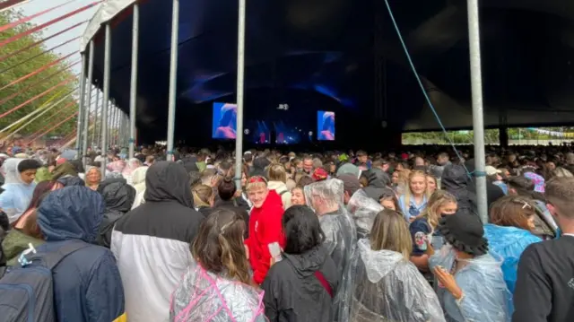 Crowd standing in front of a large stage with a tent over it. Ponchos on as rain falls.