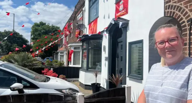 Vicky Potter, 37, who has blonde hair in a pony tail, red rimmed glasses and a grey t-shirt, stands on the doorstep of a terraced house with Liverpool FC flags and red and white bunting in the backdrop