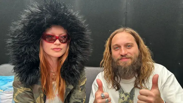 Two members of Wet Leg, one wearing a thick fur lined hoodie, and the other is giving a thumbs up.