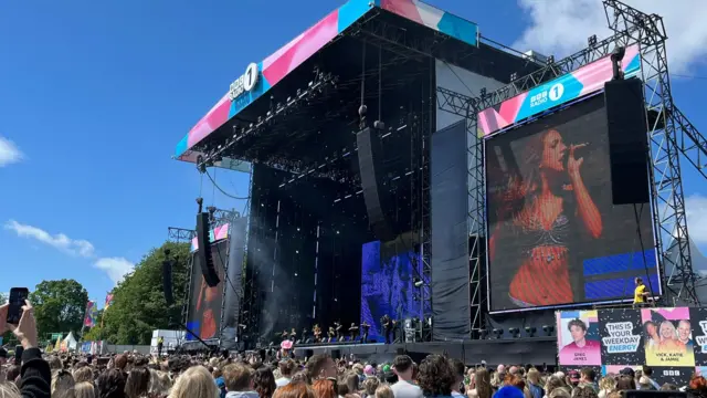 A stage with people stood around in front of it in the sun