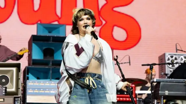 Singer Lola Young performing on stage. She is wearing jeans with a sparkly silver top and white jacket.