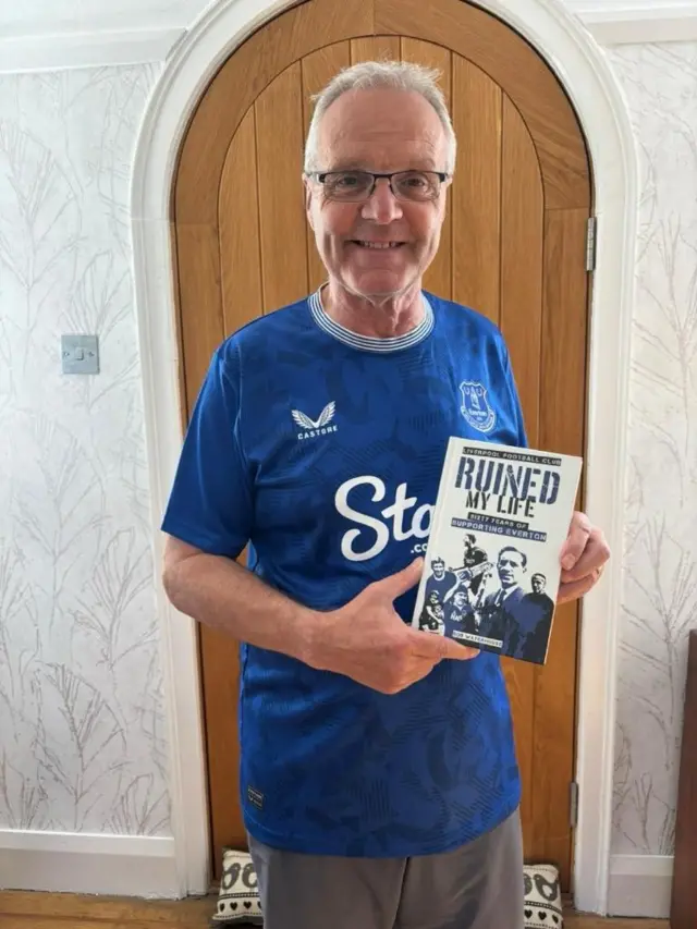 Evertonian Alan Rothwell stands holding a book called 'How the Reds ruined my life'