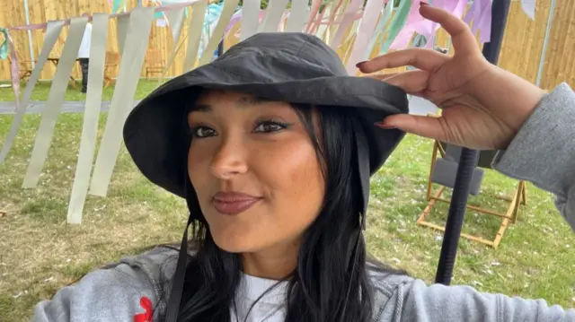 Joy Crookes posing for selfie, hand on hat and smiling, field and ribbons behind her.
