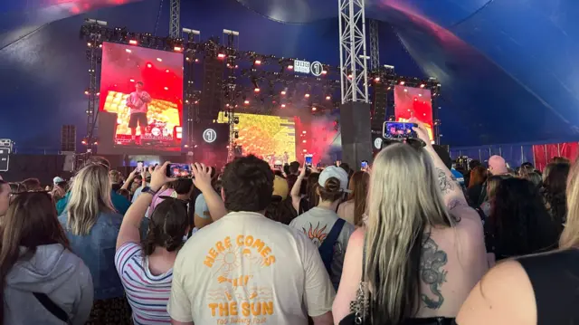 A large crowd, with people with their phones out, gathers around a stage where Hard Life is performing