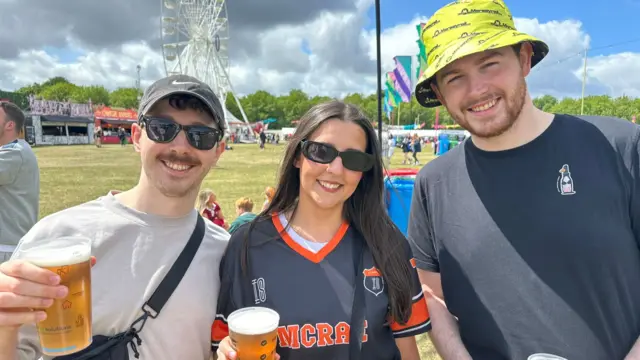 Daniel (L) in white short-sleeved shirt holds up a beer. Kate (C) in black and red American football jersey and sunglasses stands next to Tim in short-sleeved black shirt and yellow bucket hat