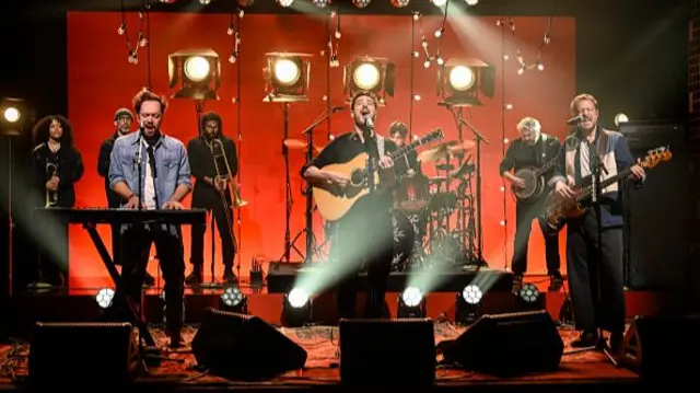Mumford & Sons performing on stage, with background musicians. There's a red background with several standing light fixtures.