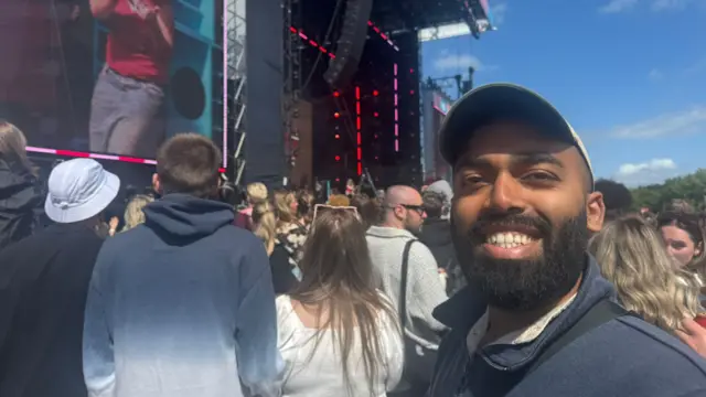 Ash smiling at the camera in front of the stage where Lola Young is performing.