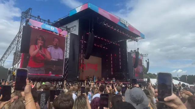 A wide shot of the stage where Lola Young is performing. There's a large crowd around it with several people with their phones out.
