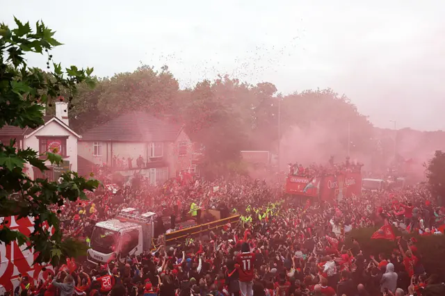 wide-angle shot of the Liverpool open-top bus parade. Red smoke from countless flares hangs in the air, enveloping the vehicles in the procession