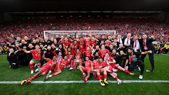 Liverpool players and staff pose in front of Kop with Premier League trophy