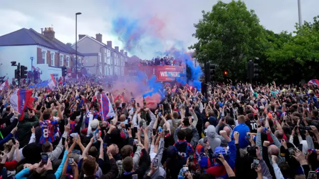 A huge crowd around a bus with smoke bombs in the  air