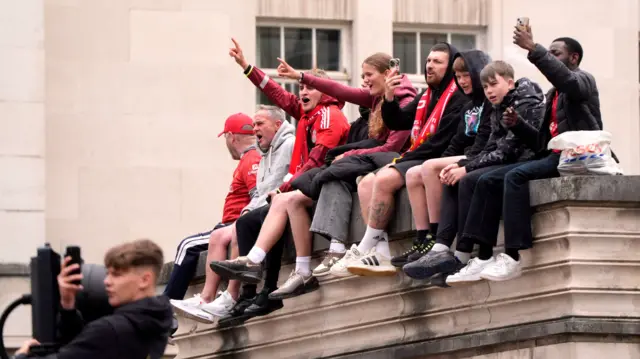 Liverpool fans in shirts and scarves sit on a high wall ahead of the parade