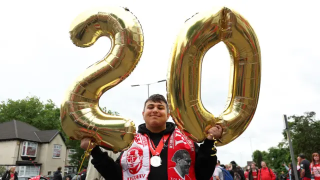 Man wearing a red Liverpool scarf holds two gold balloons, a two and a zero