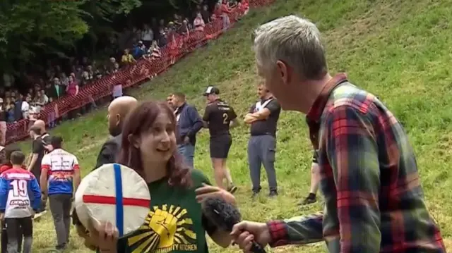 Cheese race winner in green top speaks to reporter while holding a cheese wheel