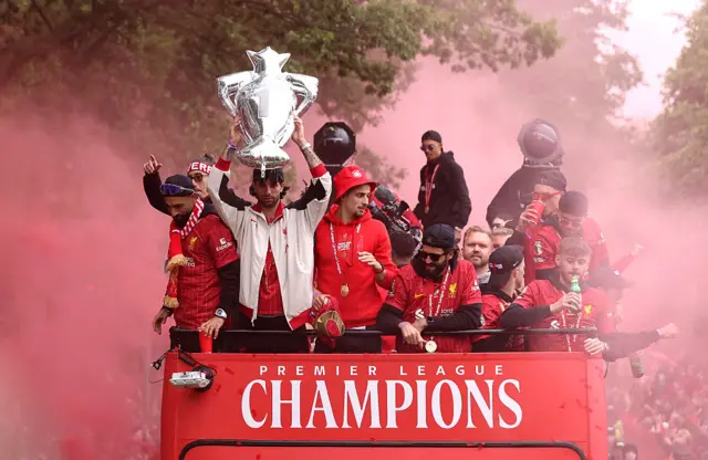 Mohamed Salah, Dominik Szoboszlai, Curtis Jones, Alisson Becker and Conor Bradley celebrate from the open-top bus with plumes of red smoke from flares all around.