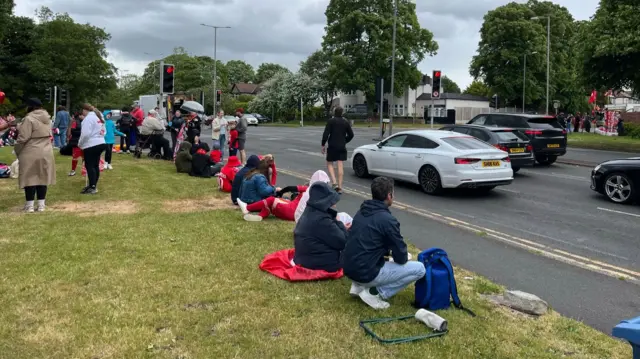 Fans on the parade route on Queens Drive sat on the grass verge of a dual carriageway