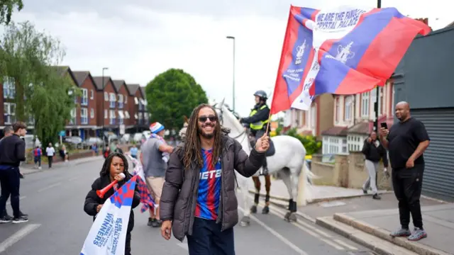 Two people walking down a street as one holds a large Palace flag.  A police horse is stood behind