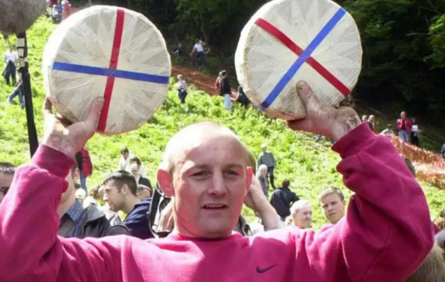 Mr Brian holds up two cheese rounds above his head. He is wearing a red nike jumper. Coopers hill can be seen behind him.