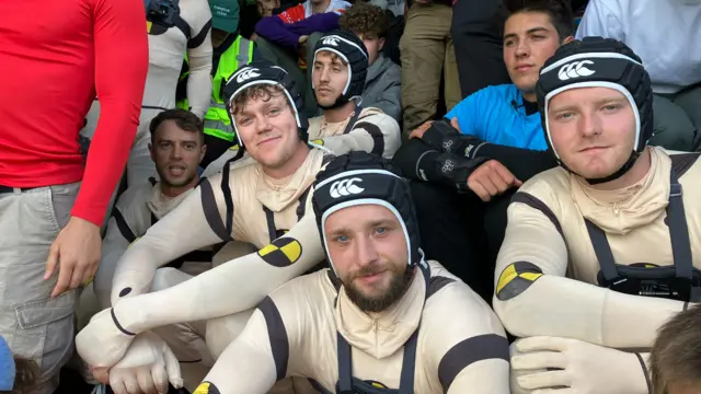 A group of men in morph suits is sitting next to each other and smiling at the camera.