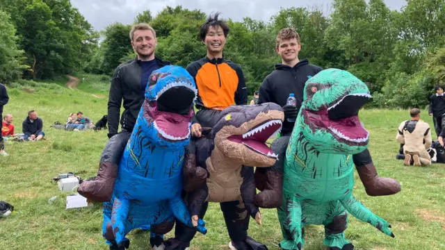 Three men smile at the camera, dressed in dinosaur suits.