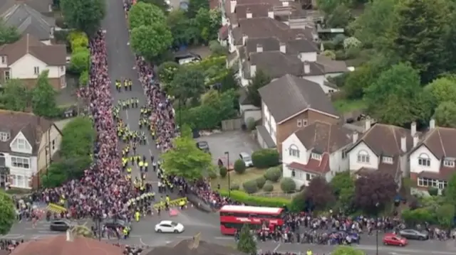 An aerial view of a street which has police in the middle and is lined by hundreds of people