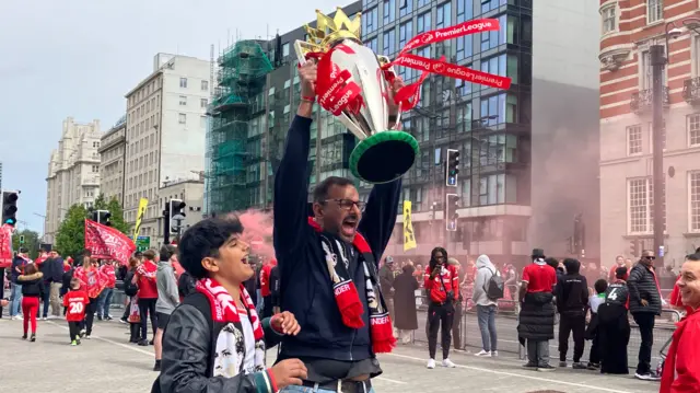 Liverpool fan Sundeep holds up a replica Premier League trophy with his son Ajay cheering on as they wait for the ope-top bus parade