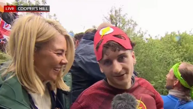 Blonde BBC reporter interviews young man with curly hair dressed as the comic book character Flash (red suit, with yellow lightning logo on white background)