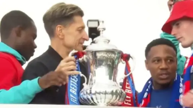 A zoomed in picture of a silver trophy with men stood around it