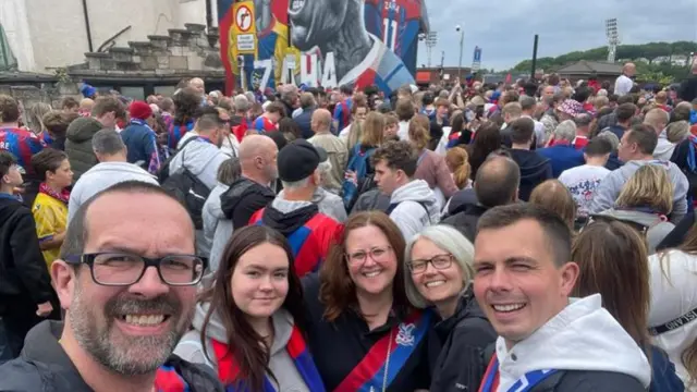 Five people, two men and three women in between them, smiling at the back of a huge crowd of Palace fans