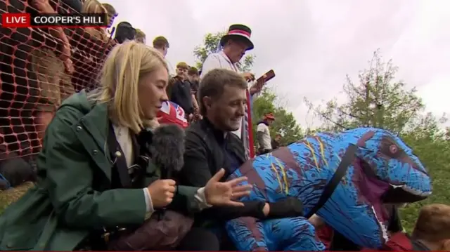 BBC reporter interviews man in dinosaur suit waiting to chase a cheese wheel downhill, the master of the cheese behind them in a top hat and white coat