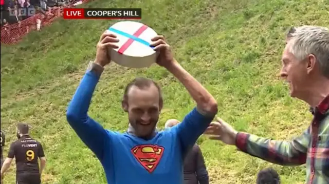 Man in a Superman suit holds up a Double Gloucester cheese wheel over his head as a man in a chequered shirt approaches him with a mic in his left hand, a grassy hill behind them