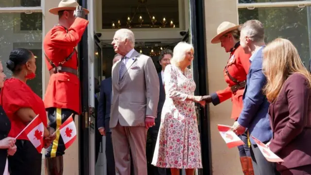Queen Camilla and King Charles on a red carpet, Camilla shaking hands with Canadian mounties holding Canadian flags