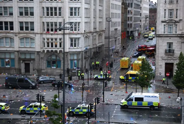 A littered street is empty, with only police cars and ambulances parked and a few officers stood around