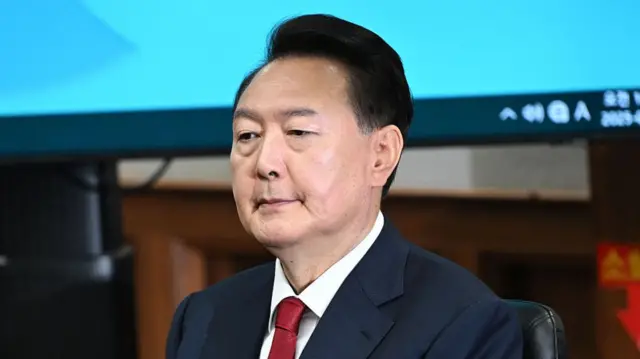Yoon Suk Yeol in a navy suit and red tie, he looks solemn as he sits on a leather chair
