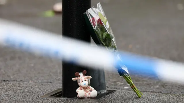 Flowers wrapped in cellophane and a stuffed giraffe toy lie on the ground near the incident site. In front of it, blurred, is police blue and white tape