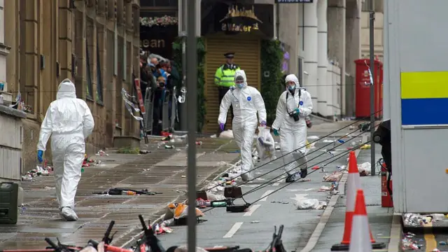 Two forensics workers walks down a road, one is carrying a bag and the other a camera. Another forensics worker walks the other. A police officer can be seen in the distance.