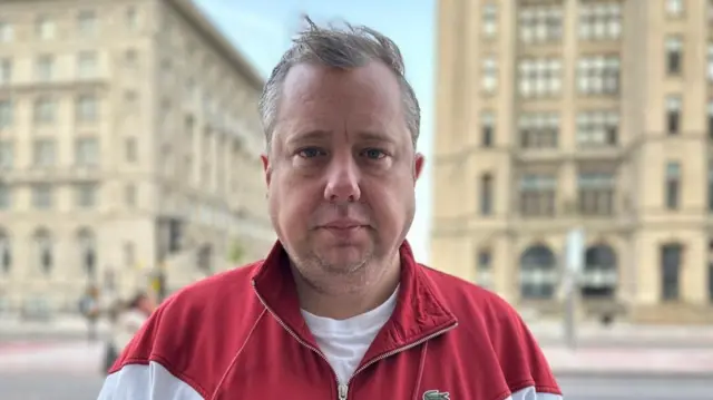 A white man in a red jacket stares blankly into the camera on a street in Liverpool.